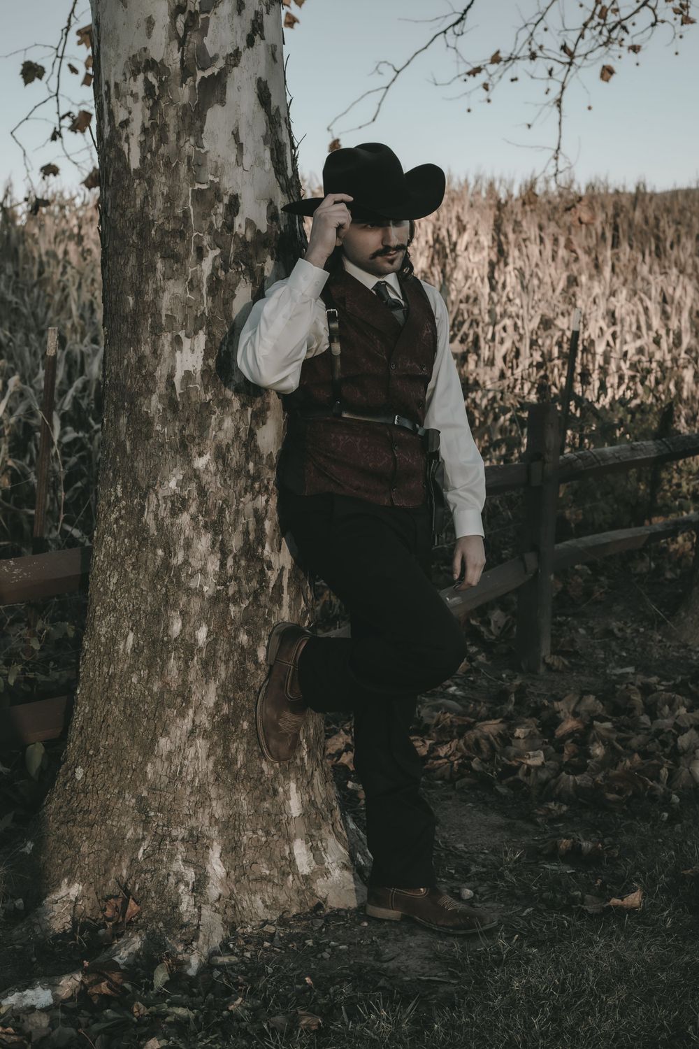 Man in Western cowboy vest and hat leaning against tree by rustic fence in autumn setting.