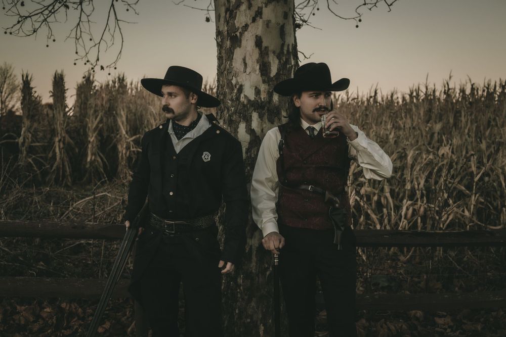 Two men in vintage Western cowboy costumes standing by tree near cornfield at dusk.