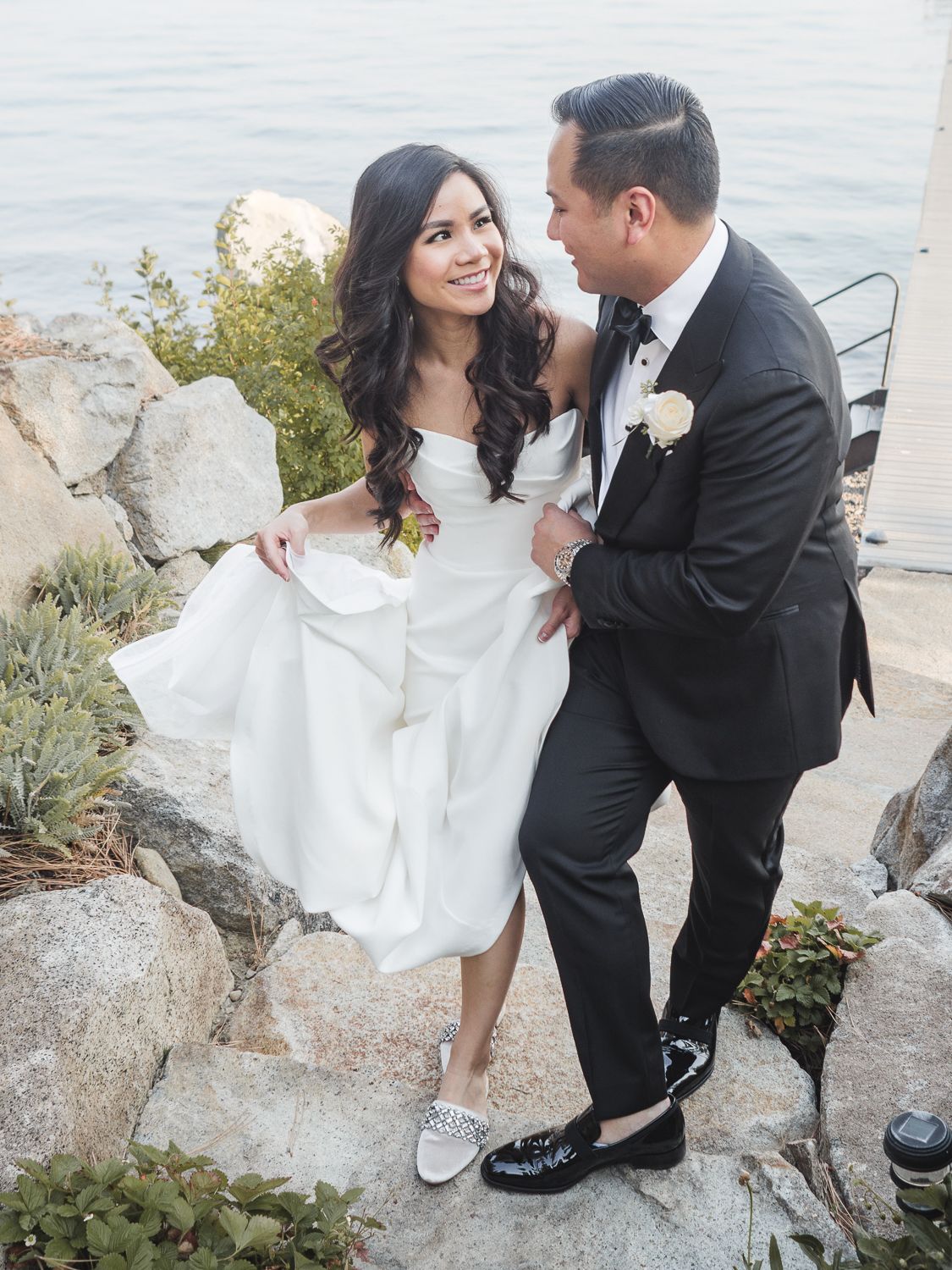 A bride in a white gown and groom in black tuxedo pose together on rocky waterfront steps wearing matching sneakers.
