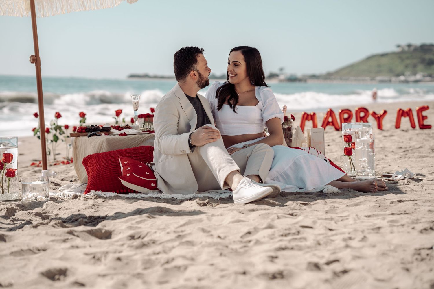 Couple enjoying picnic on beach with marry me sign and roses in background.