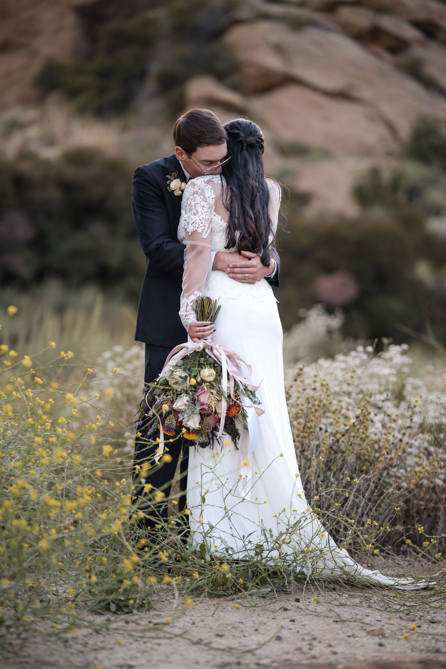 A romantic wedding photo of a couple embracing outdoors with a rustic desert backdrop and wildflowers.