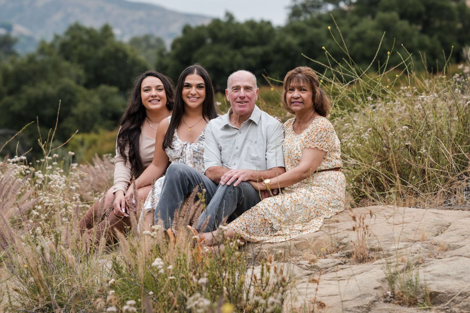 A family group sits together on a natural hillside with mountains in the background.