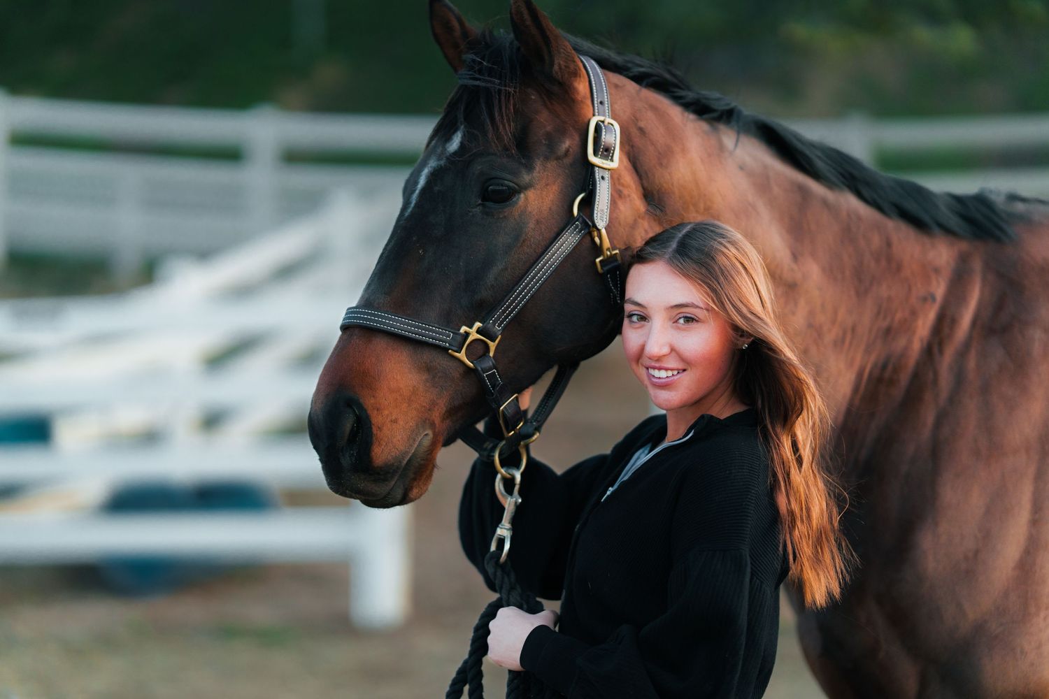 Young girl in black outfit stands next to brown horse with bridle.