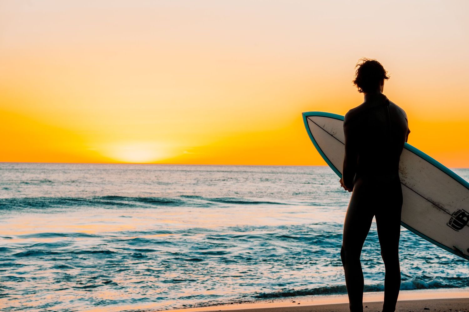 Silhouette of surfer holding board against vibrant orange sunset over ocean.