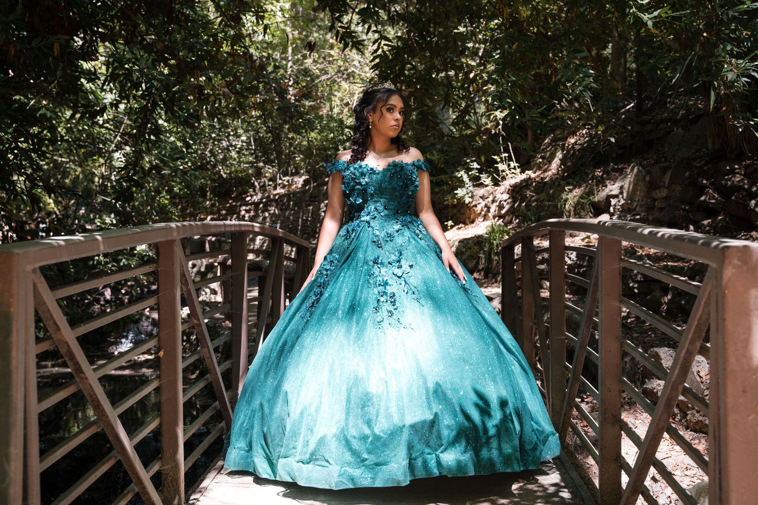 A stunning turquoise quinceañera dress sparkles on a wooden bridge surrounded by lush greenery.
