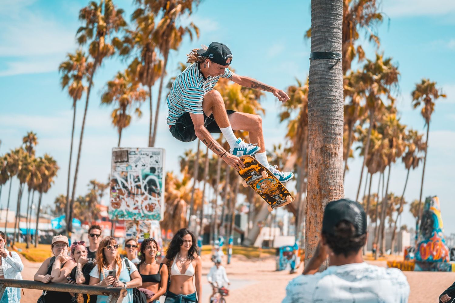 A skateboarder performs an impressive jump at a beachside skate park with palm trees in the background.