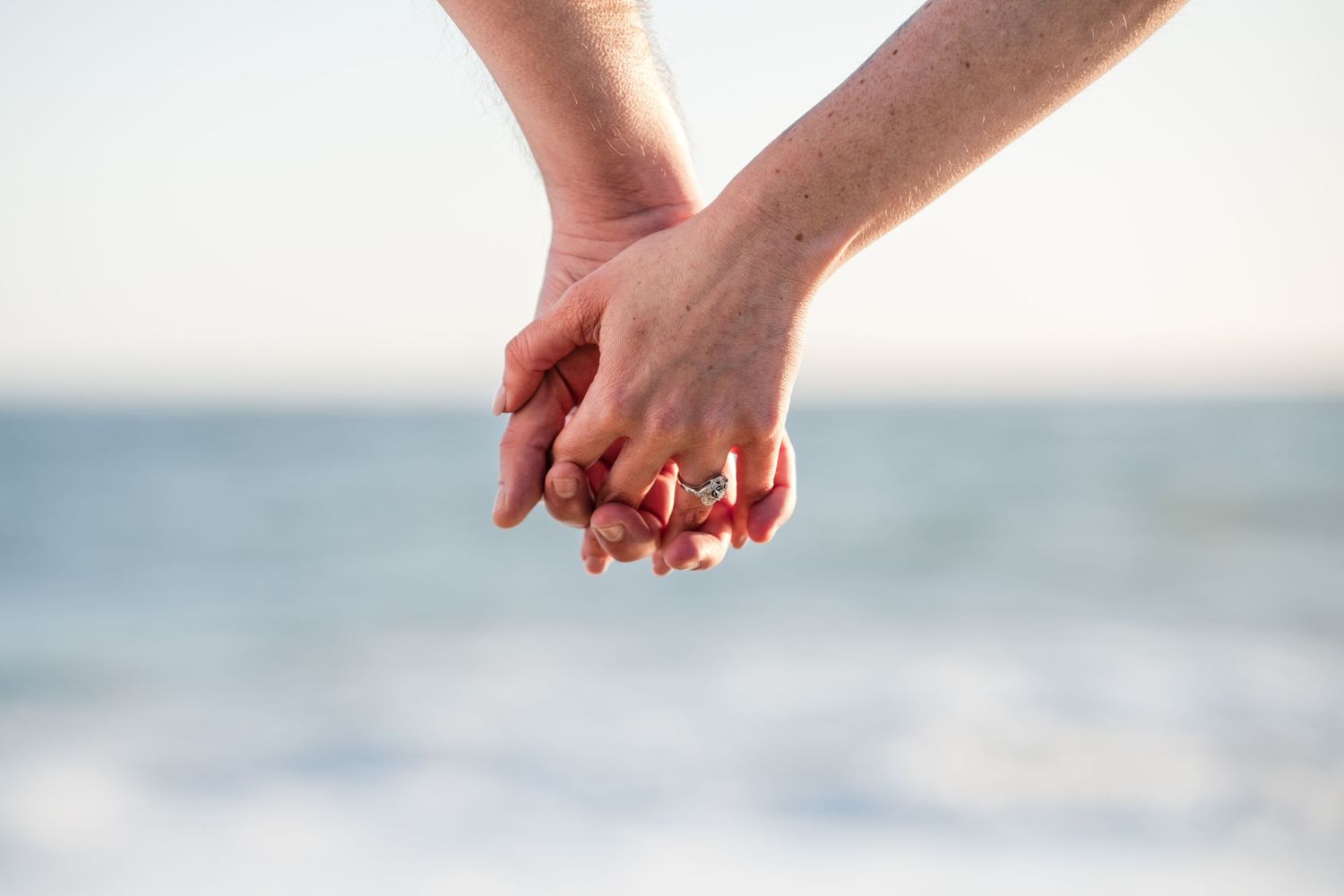 Two hands clasped together romantically against a blurry ocean background at sunset.