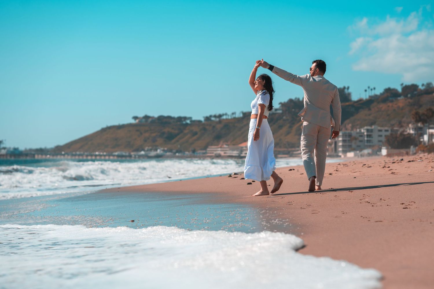 Couple dancing playfully together on beach with waves and cliffs behind them.