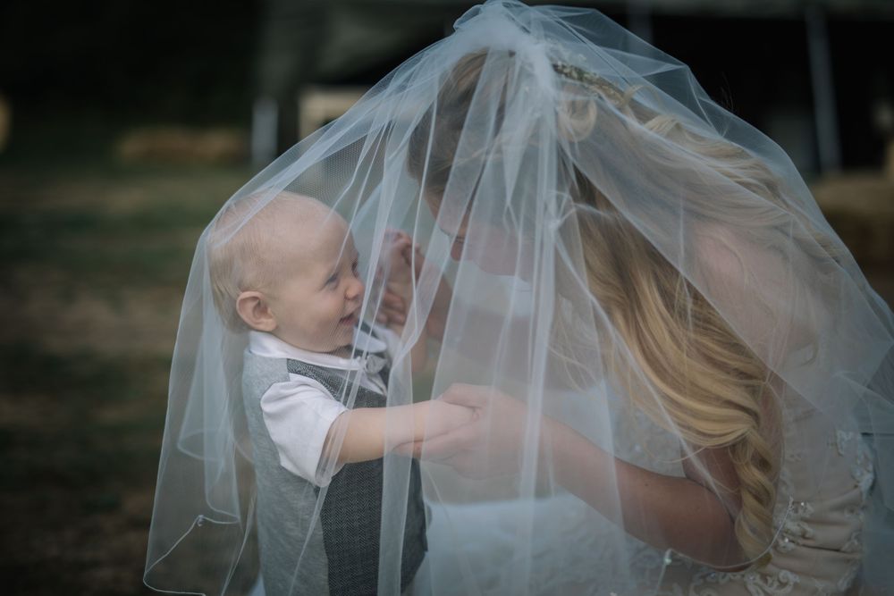 Sweet candid moment of bride with baby playing under her veil during outdoor wedding photos.