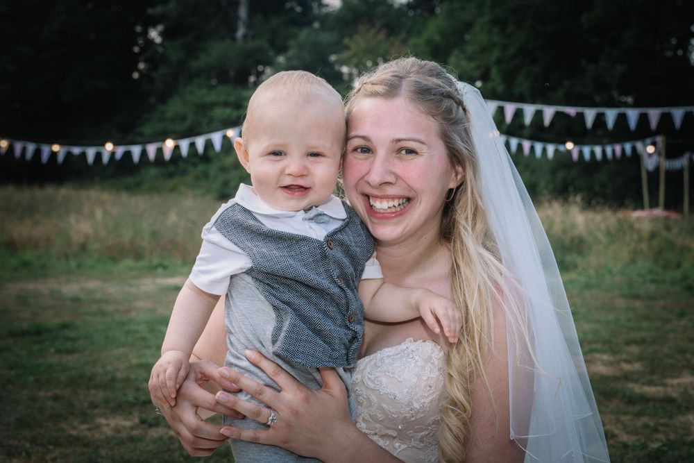 A joyful bride in her wedding dress holding a smiling baby outdoors with bunting in the background.