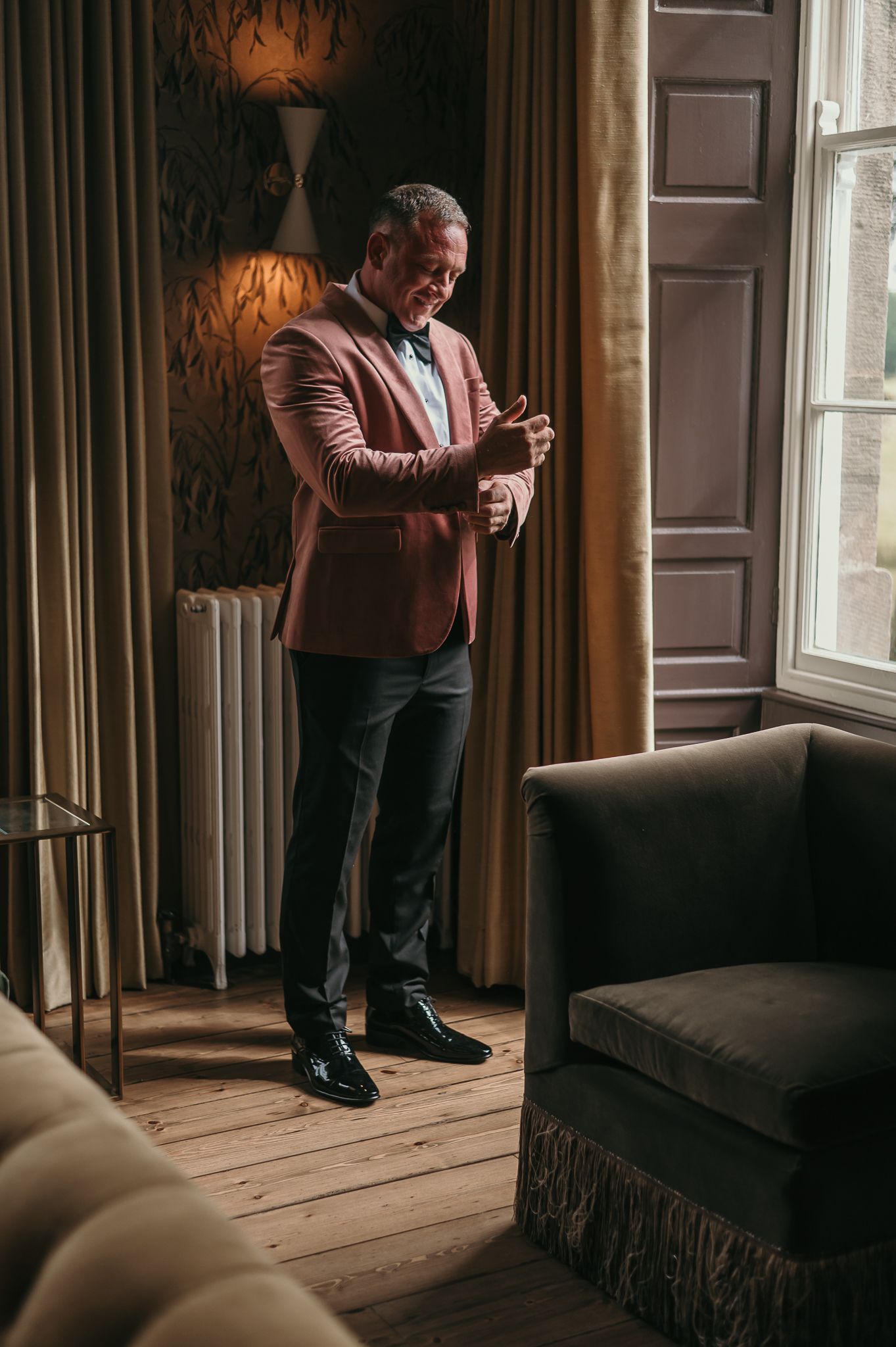 A person in a burgundy blazer adjusts cufflinks while standing by a window in an elegant room.