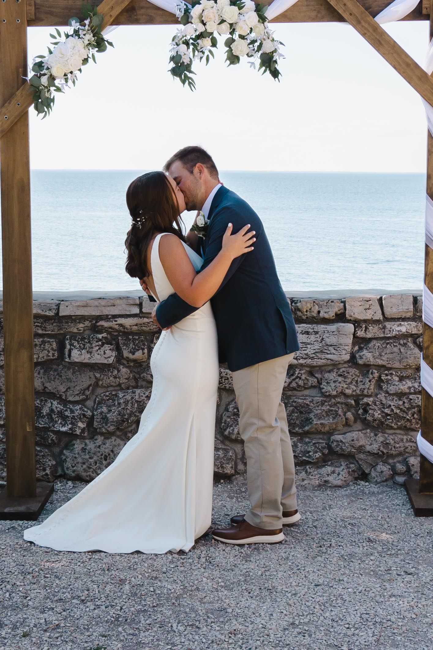 Couple sharing romantic moment under floral arch with ocean backdrop.