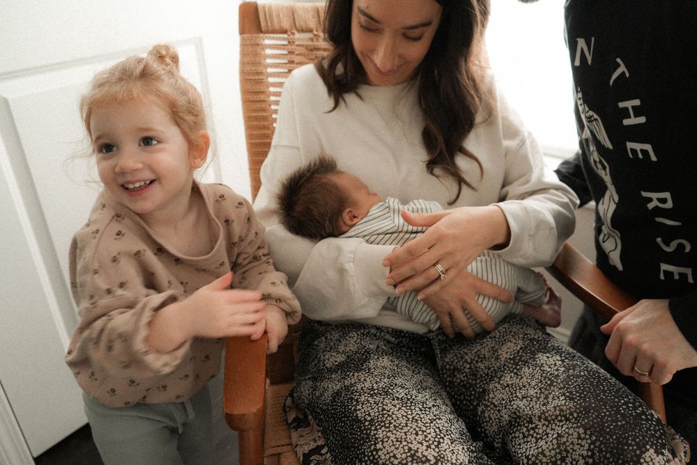 Mother breastfeeding newborn baby while smiling toddler daughter stands beside her in bright room.
