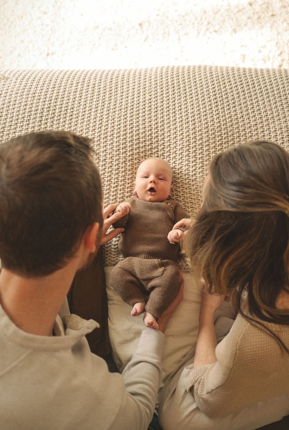 New parents looking down at newborn baby in brown knit outfit lying on textured beige sofa.