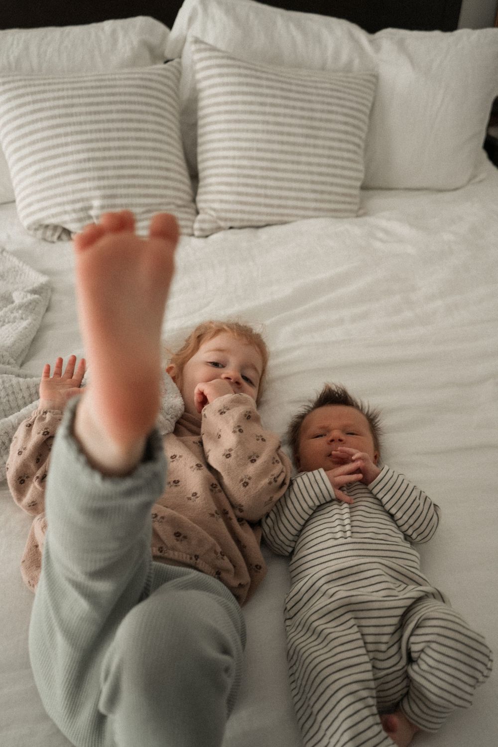 Toddler and newborn baby lying together on white bed with striped pillows, sibling raising foot.