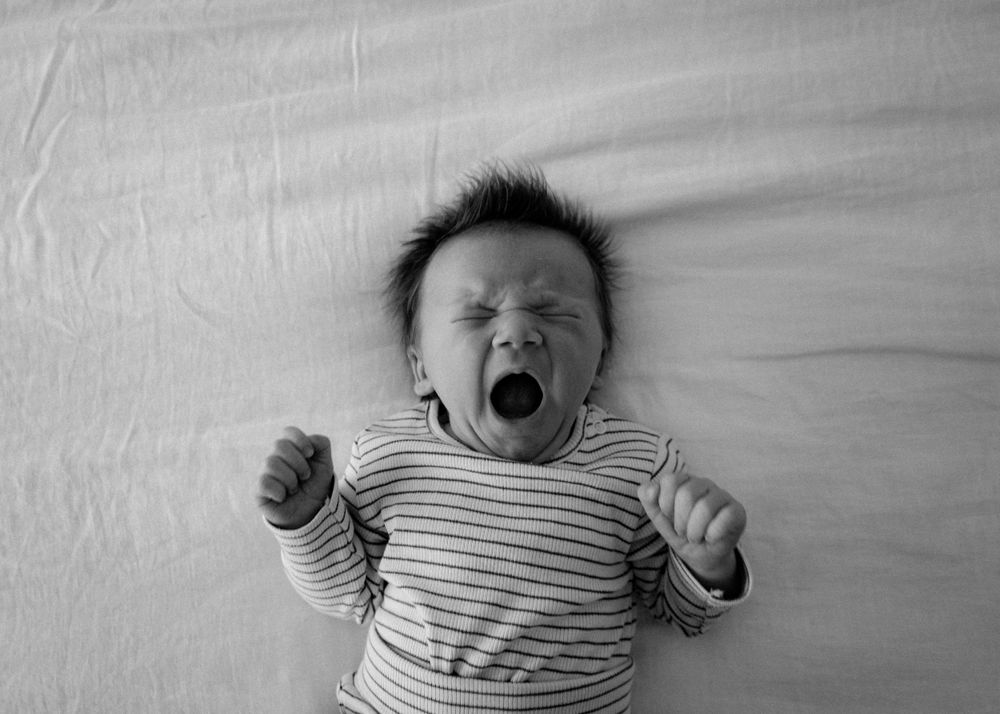 Black and white close-up of yawning newborn baby in striped onesie lying on white blanket.