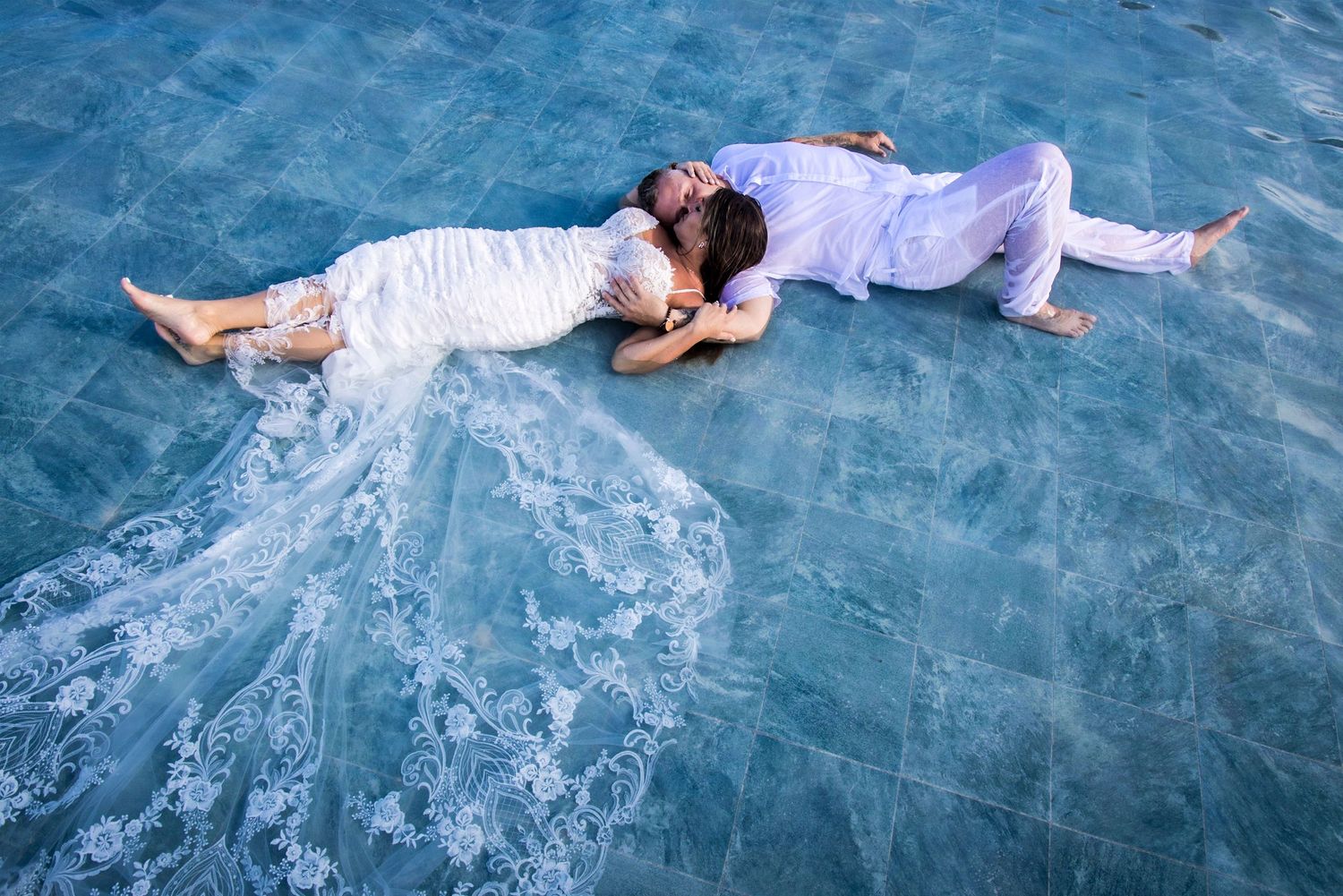 A couple in white clothing lying on crystal clear blue water in a dreamy beach setting.