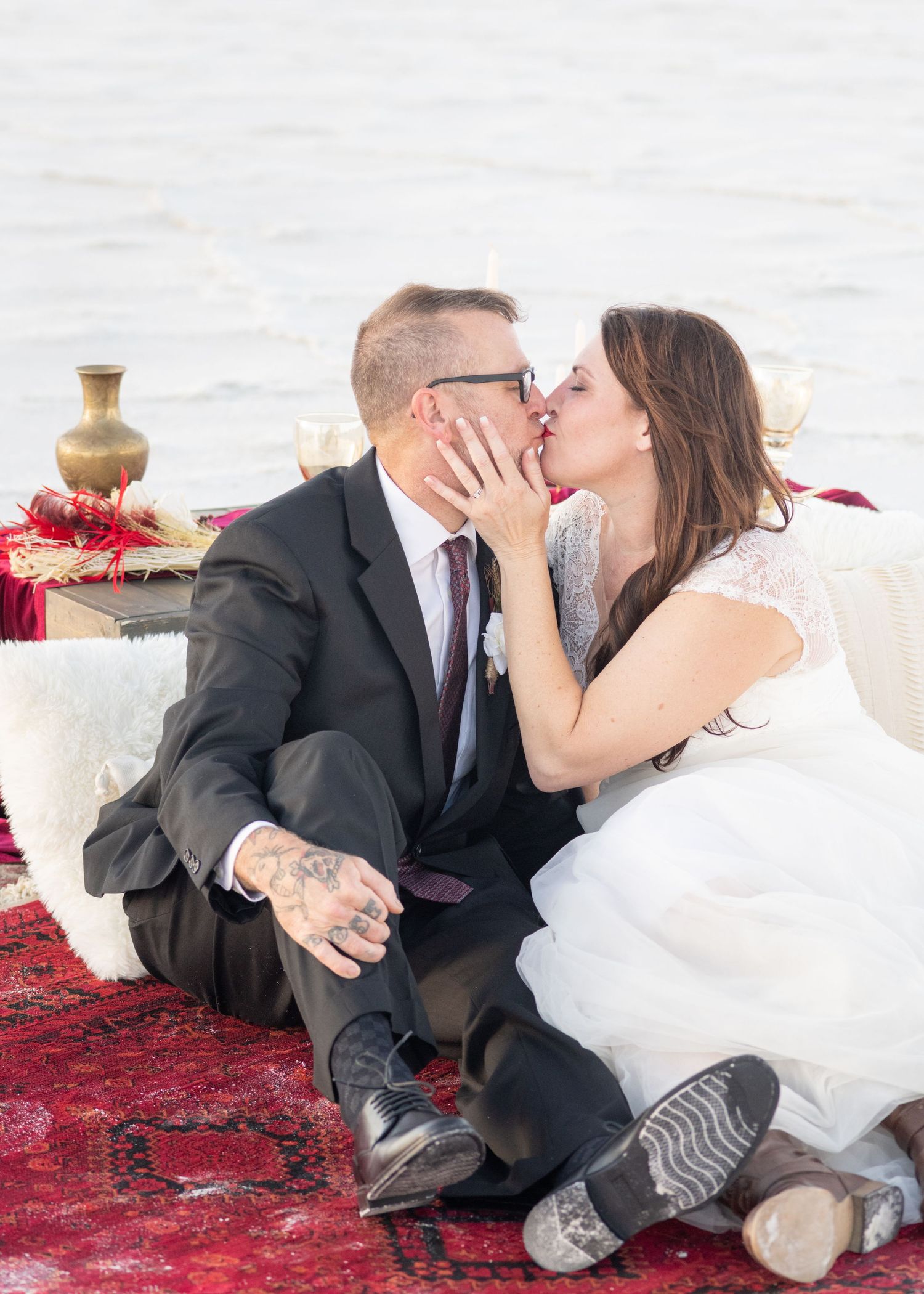 A romantic wedding photo showing a couple sharing a kiss while sitting on a red rug with decorative elements.