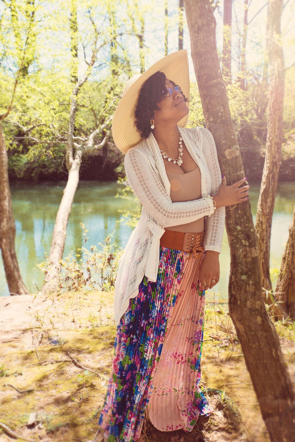 Woman in flowing skirt and sunhat poses gracefully by a stream in dappled sunlight.