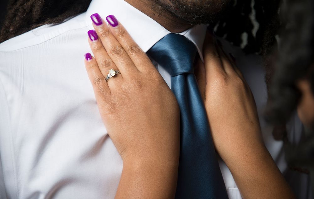 Close-up of hands with purple nails and engagement ring resting on white shirt and blue tie.