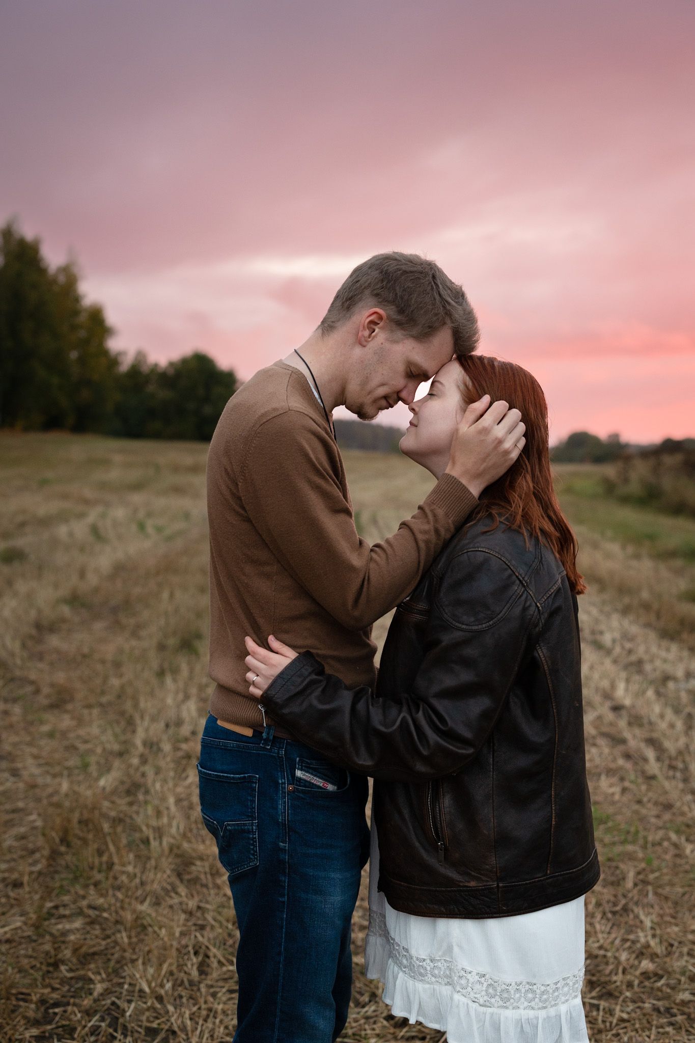 Man and woman standing on a field leaning their foreheads on each other, with closed eyes. Pink sunset background.