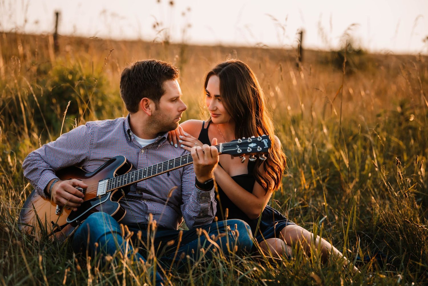 A romantic couple sits in a golden field at sunset playing acoustic guitars together.