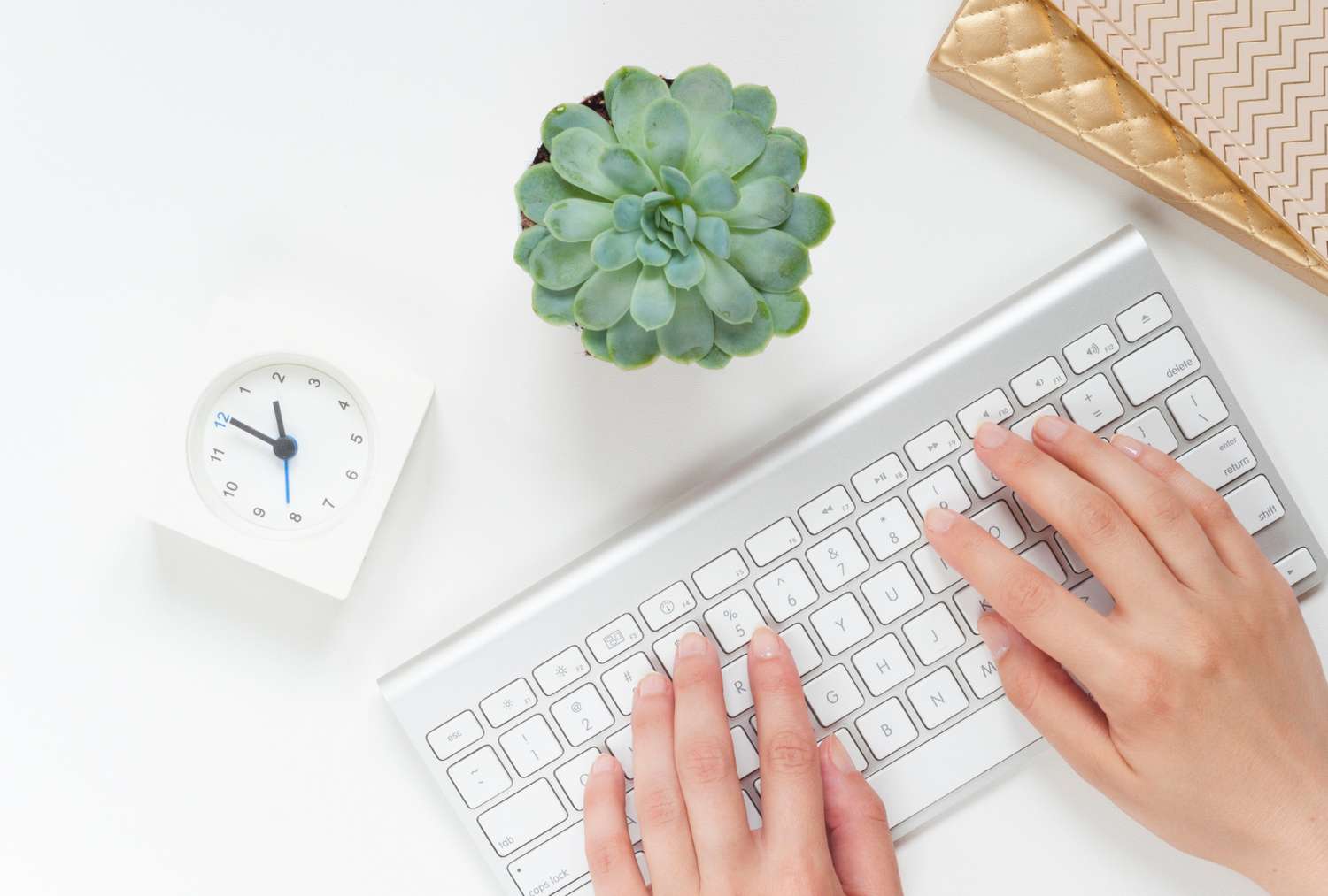 Close up photo of an apple keyboard with hands typing, a clock, a plant and some notebooks