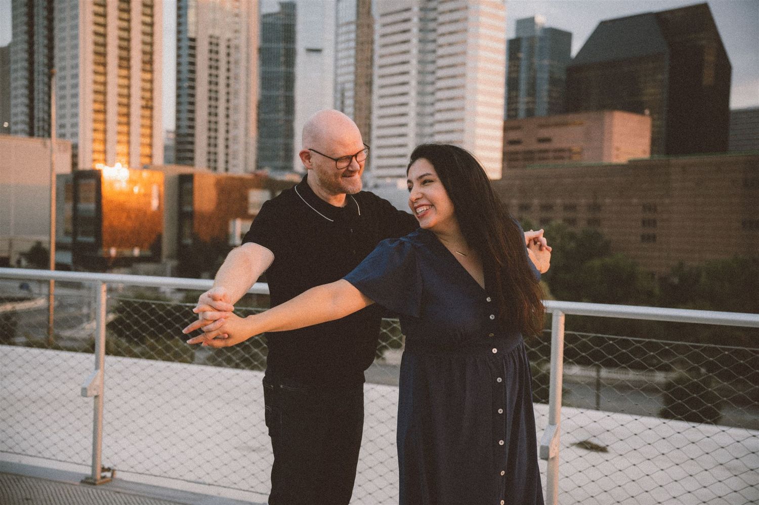 Couple dancing and laughing together on a city rooftop with skyscrapers in the background.