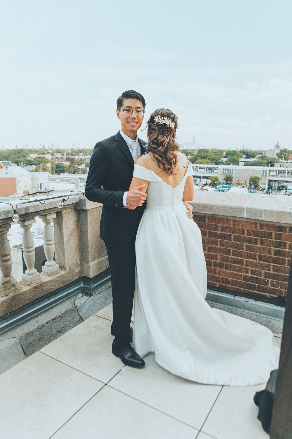 Couple in wedding attire standing on a rooftop terrace with cityscape in background.