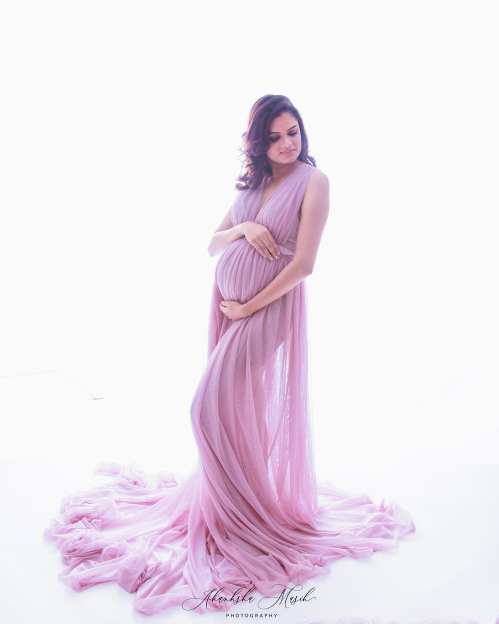 Expectant mother in flowing pink chiffon dress poses elegantly in soft white studio setting.