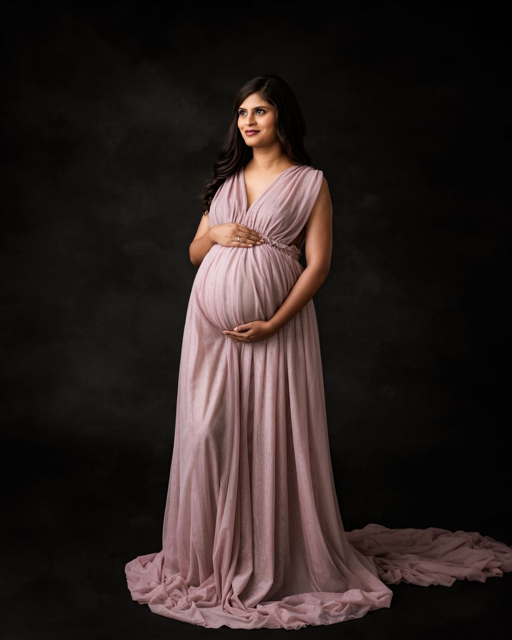Elegant maternity portrait with a flowing mauve gown against a dark background.