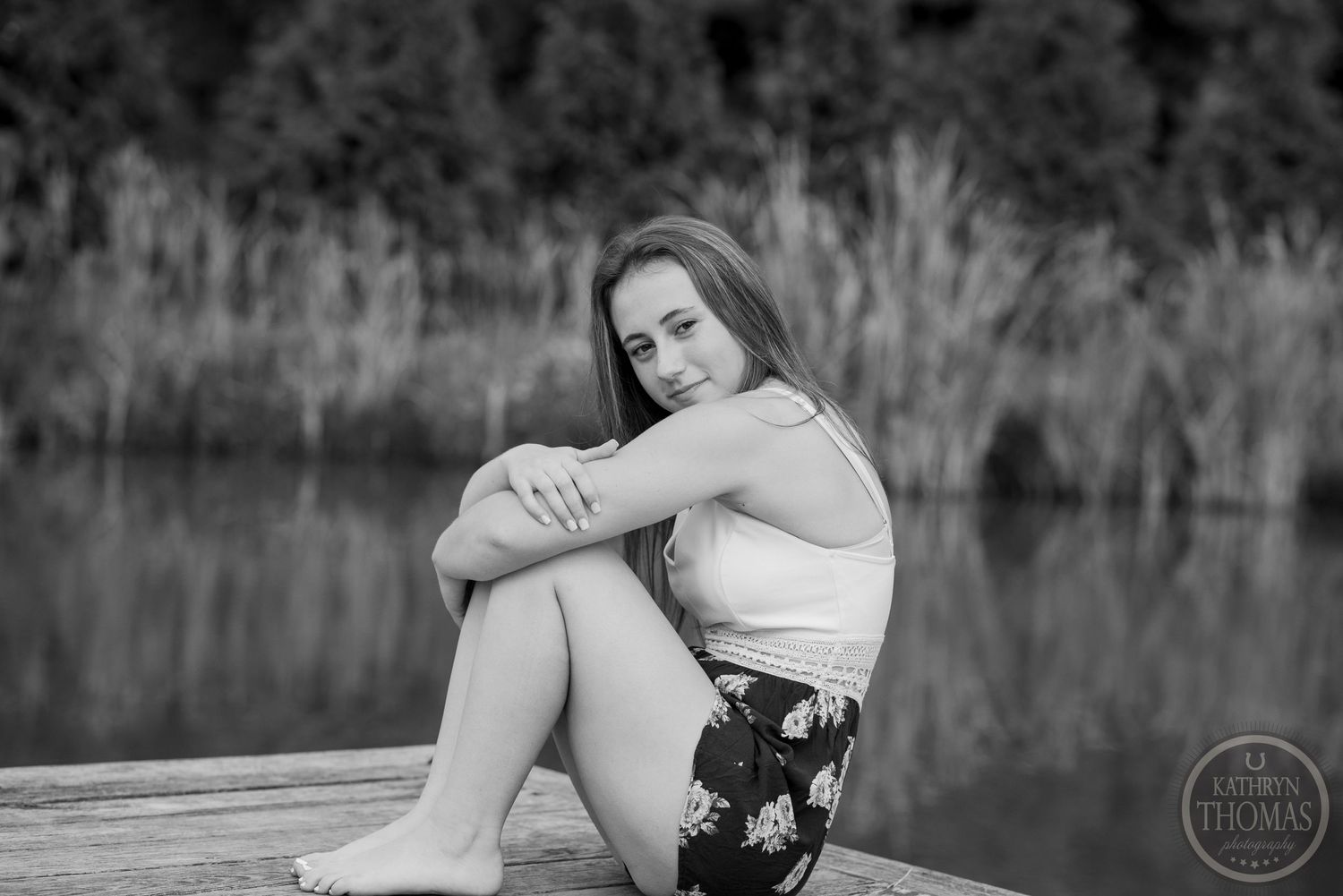 Black and white portrait of someone sitting on a wooden dock by a tranquil lake wearing floral shorts.