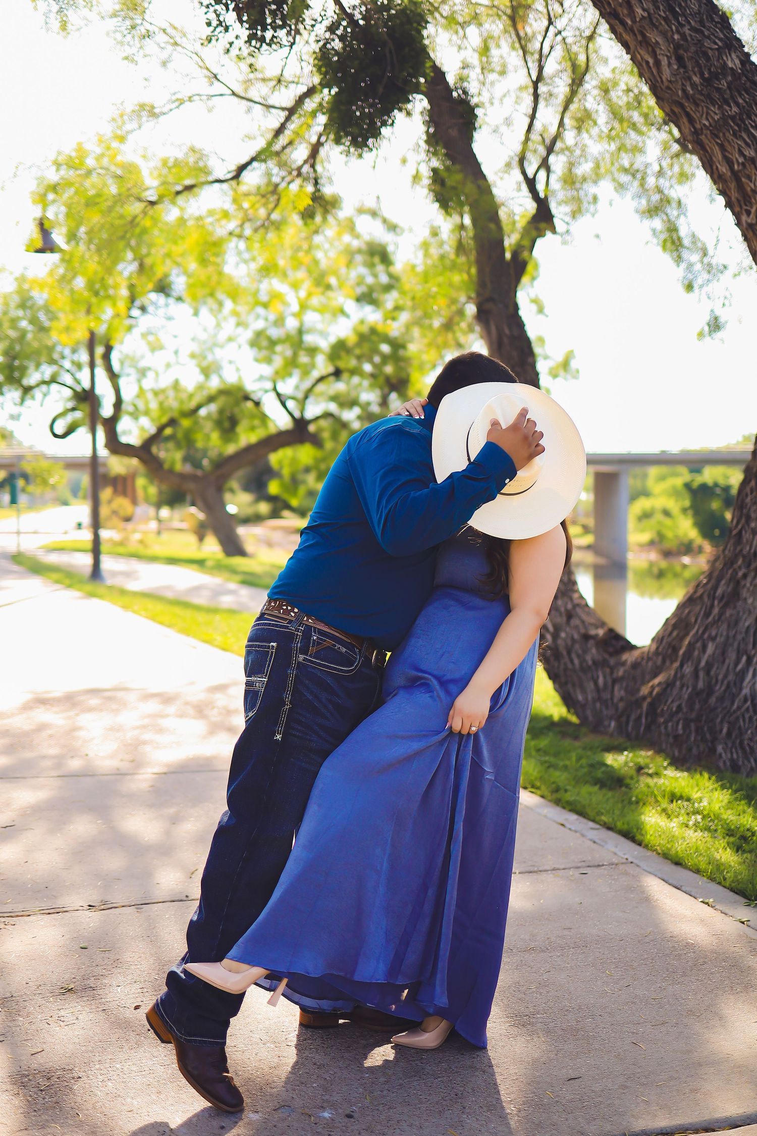 A romantic couple shares a dance pose under trees on a sunny sidewalk.