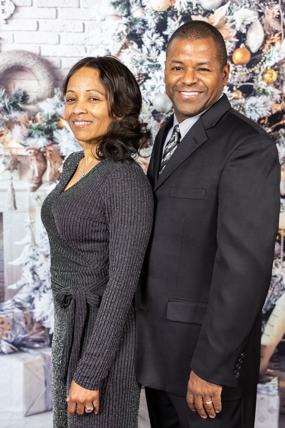 Couple in formal attire poses in front of flocked Christmas tree and white brick wall.