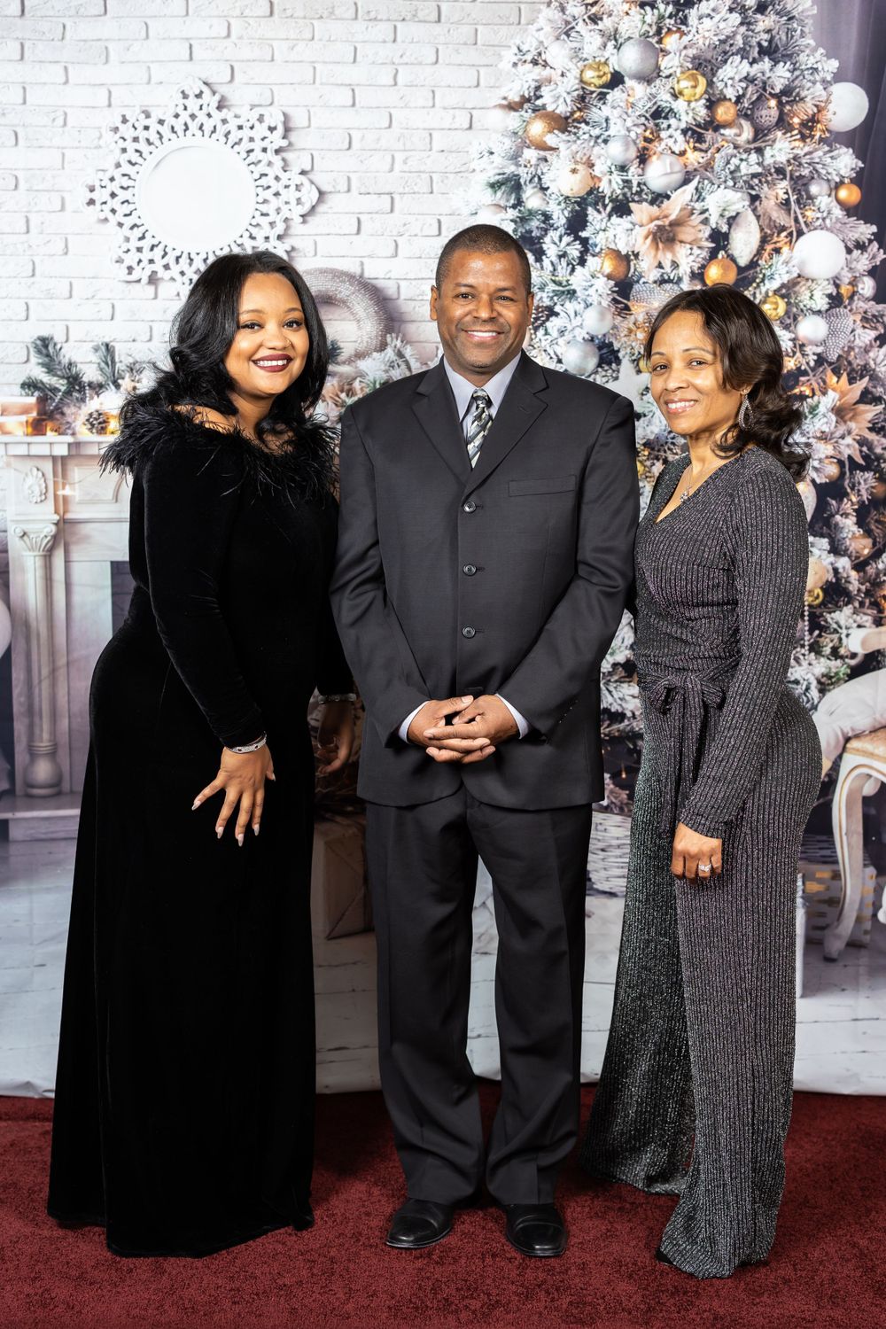 Three people in formal wear pose for holiday portrait with decorated Christmas tree backdrop.