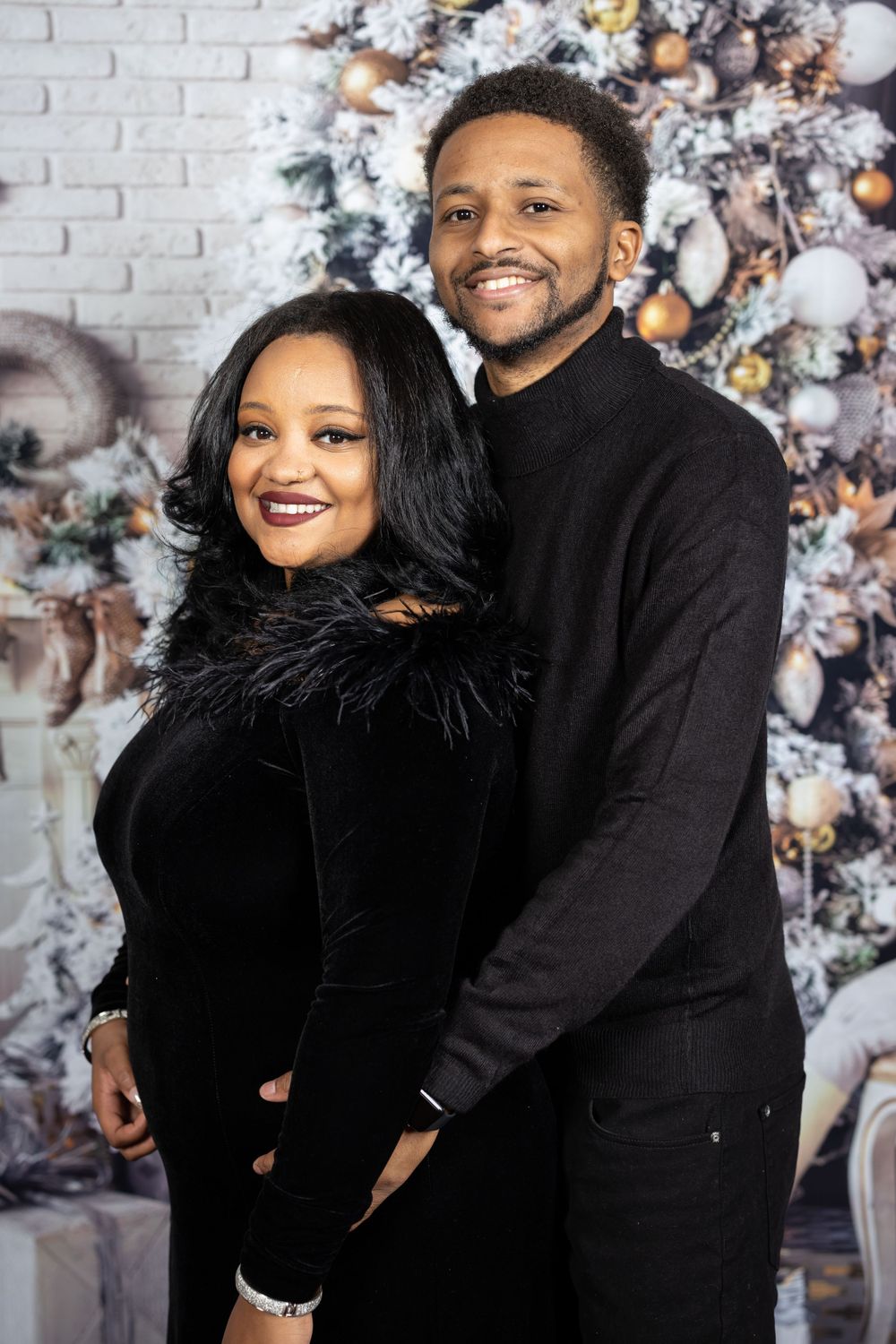Couple in black formal wear poses together in front of flocked Christmas tree.