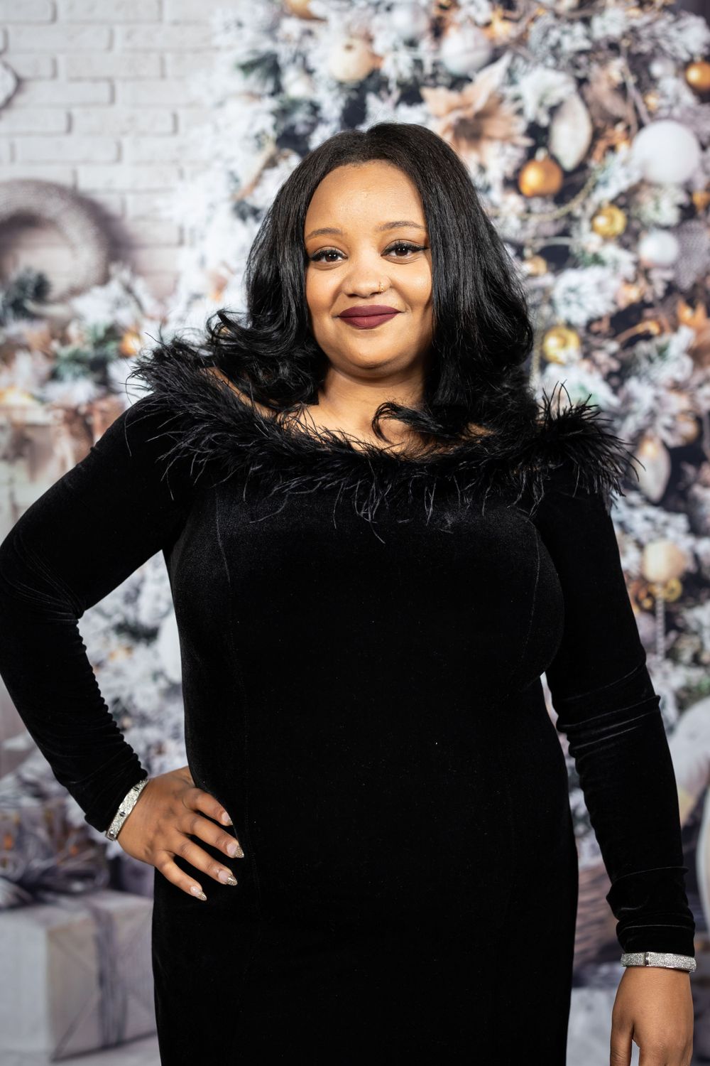 Elegant woman in black velvet dress with feather trim poses against decorated holiday backdrop.