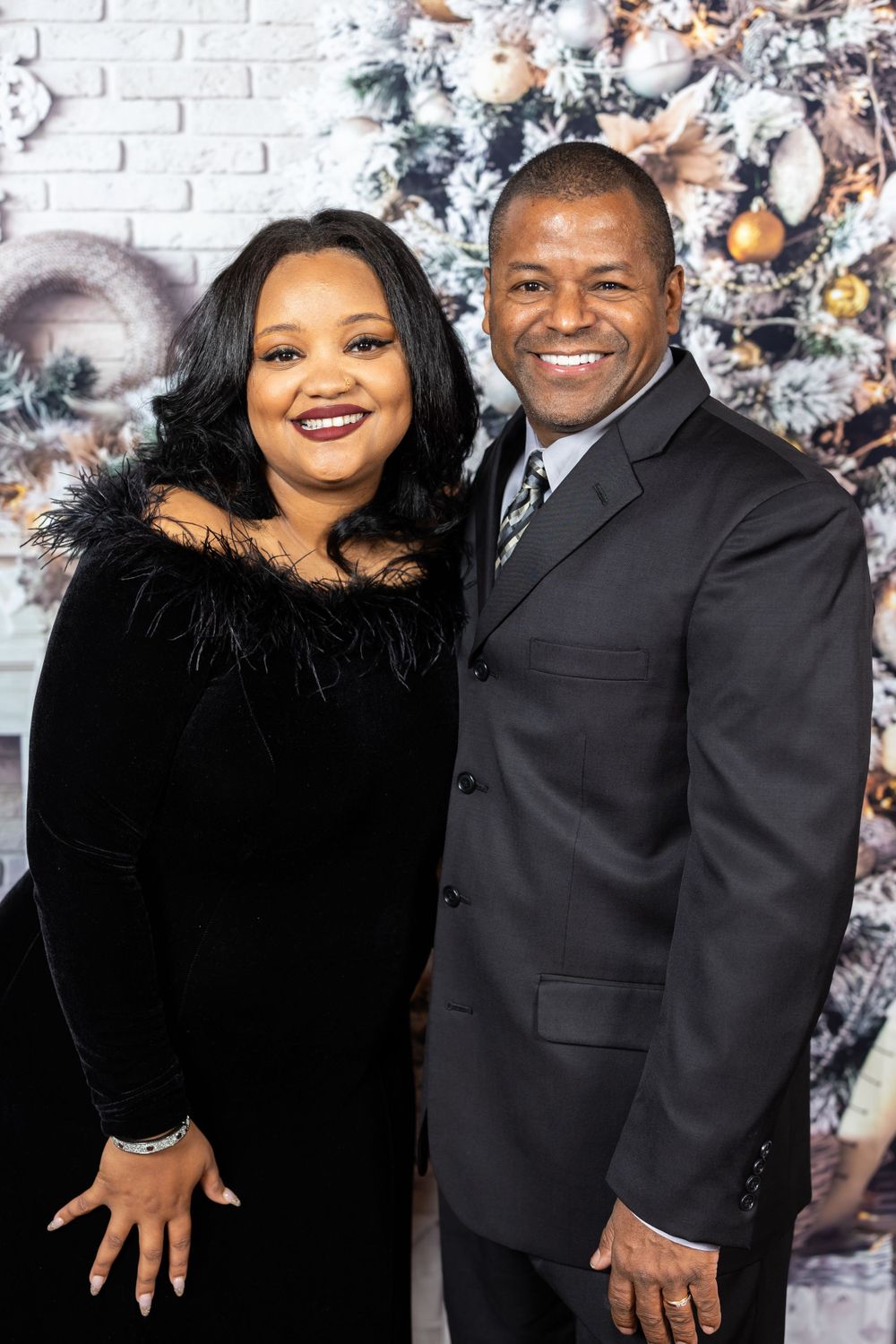 Couple in formal attire smiles in front of decorated Christmas tree and white brick wall.