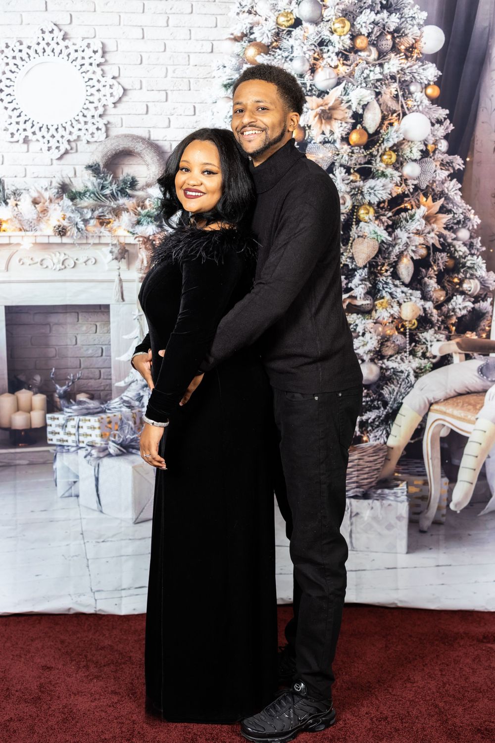Couple in matching black outfits embrace in front of a decorated Christmas tree and white backdrop.