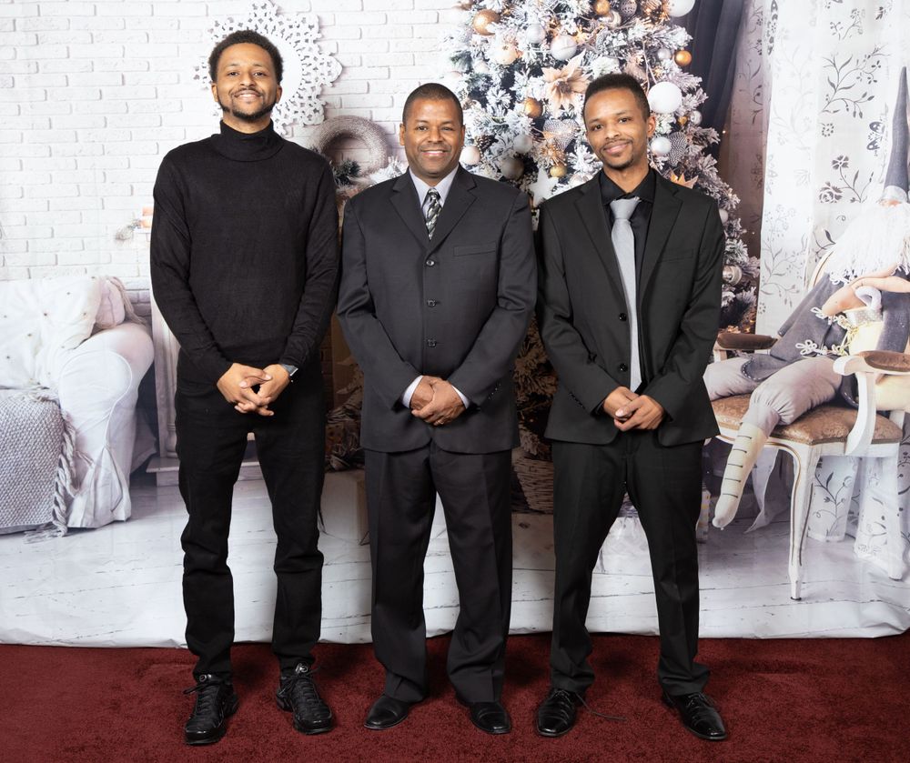 Three people in dark formal attire pose in front of a decorated Christmas tree backdrop.