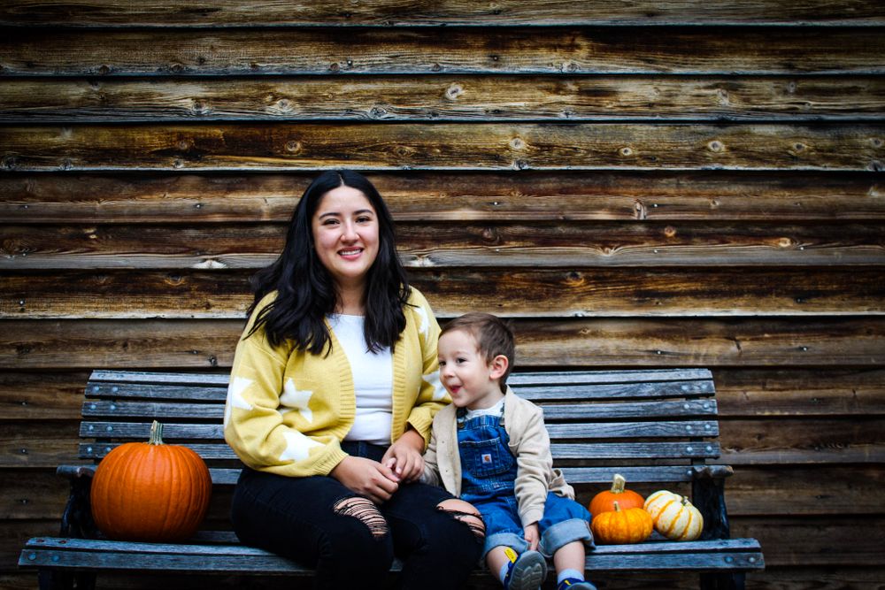 Two people sit together on a wooden bench with pumpkins against rustic wood paneling.
