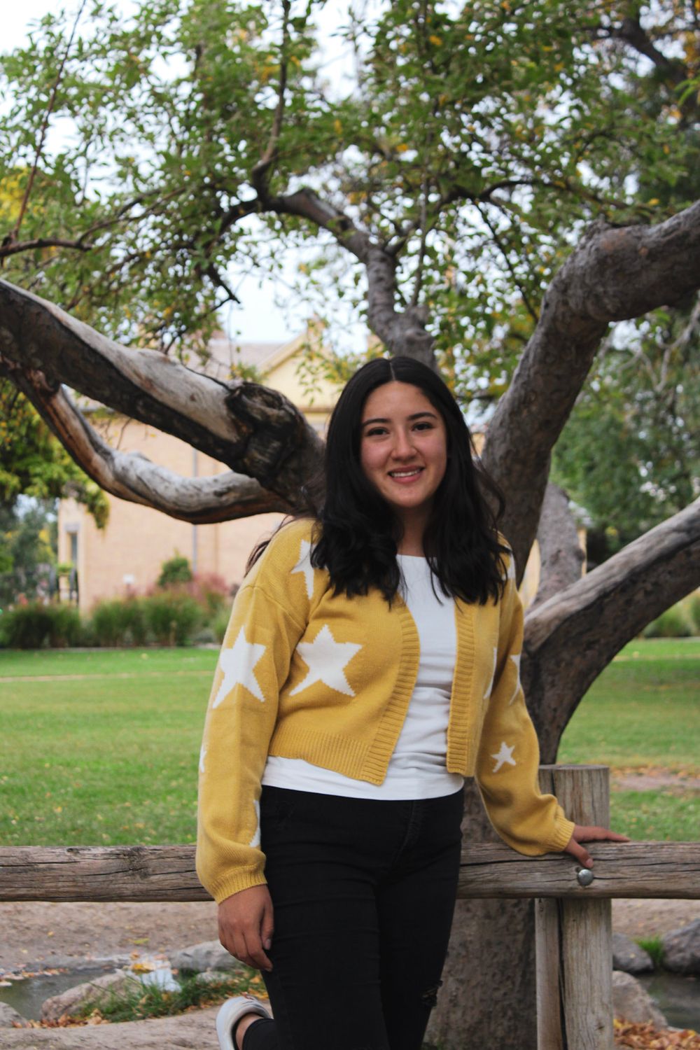 Person in yellow star sweater smiles while leaning against tree trunk outdoors.
