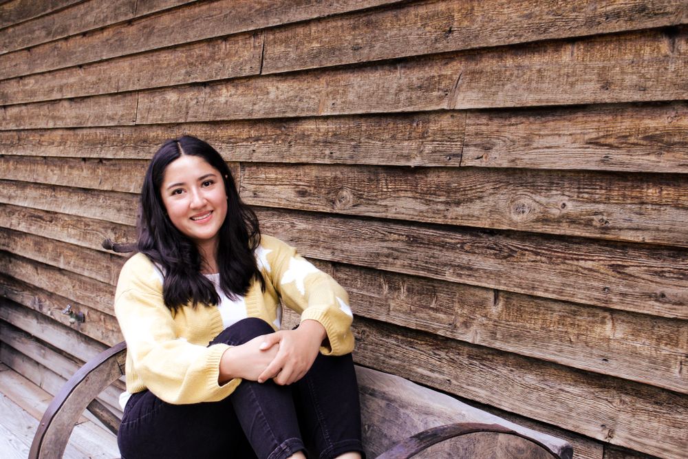 Person in yellow sweater smiles while sitting on bench against rustic wood wall.