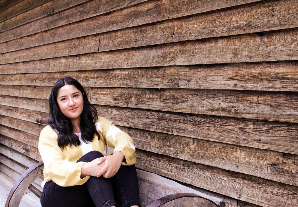 Person in yellow cardigan sitting on bench against weathered wooden wall background.