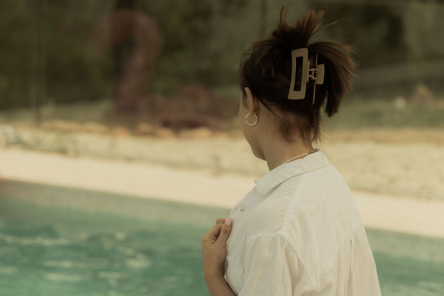 A person in white gazes thoughtfully at a poolside scene on a sunny day.