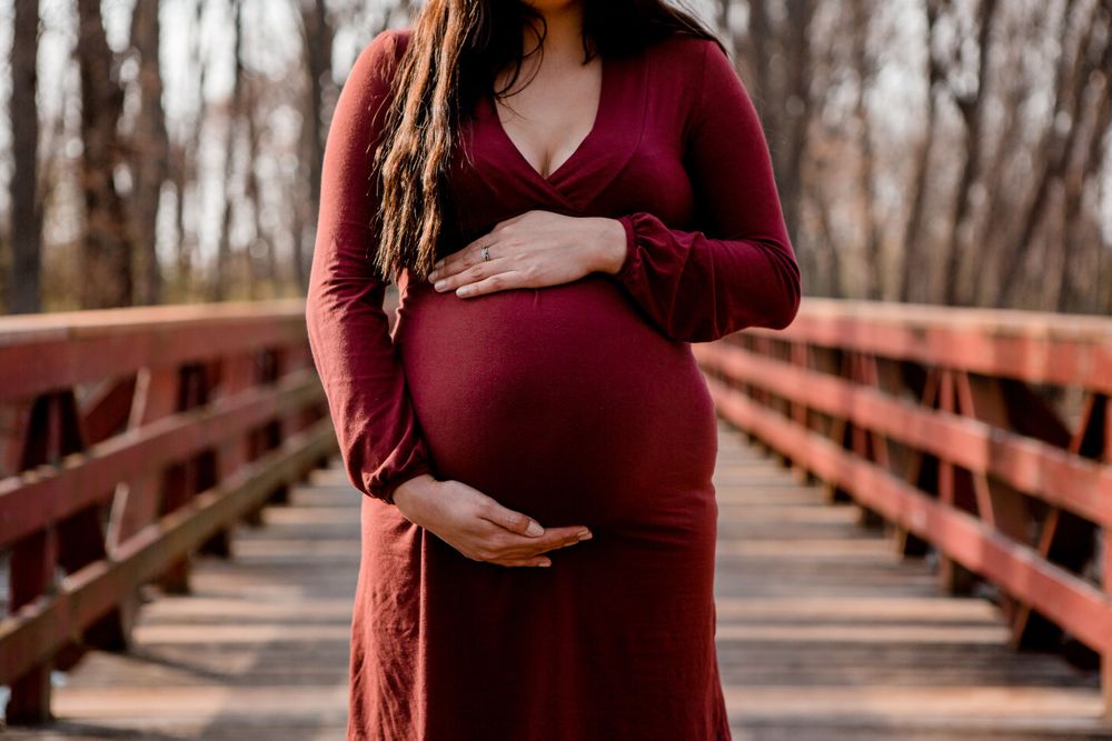 A pregnant woman in a burgundy dress stands on a wooden bridge during autumn.