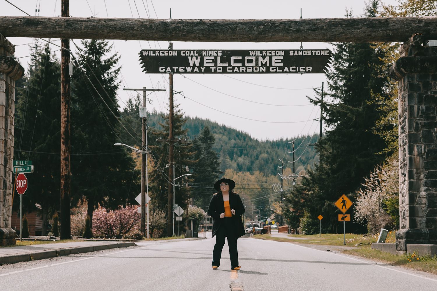 A woman stands in the middle of the street underneath a wooden sign that says Welcome