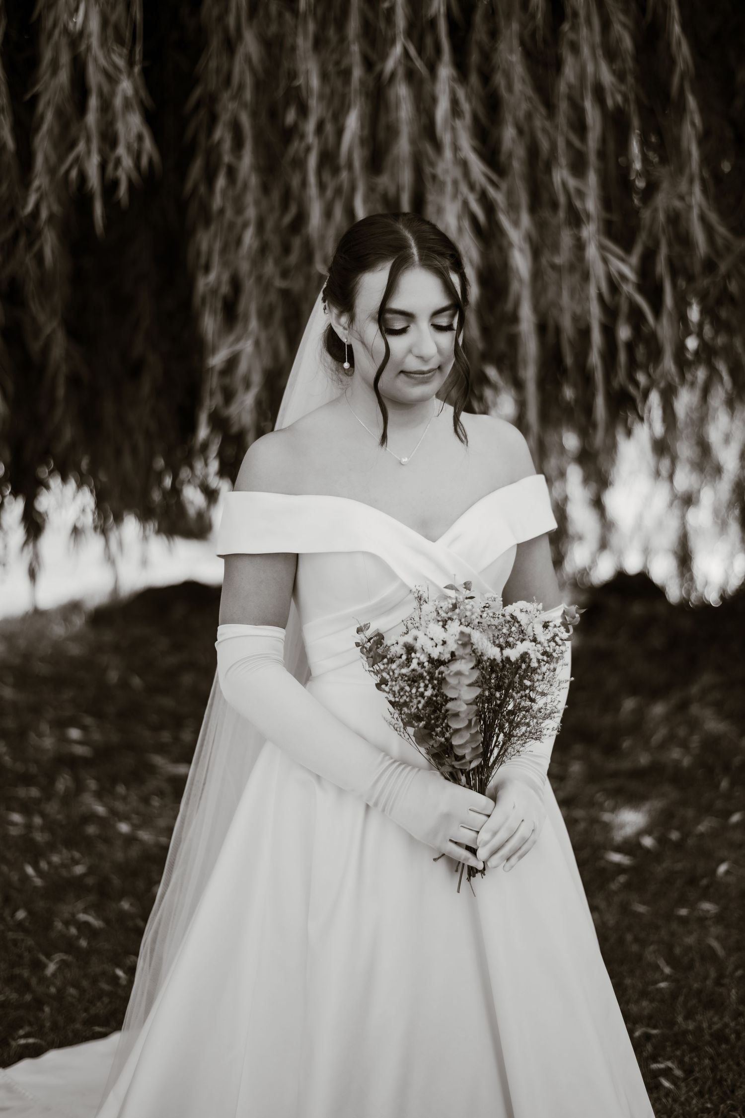 A bride in an elegant white off-shoulder gown holds a delicate bouquet against a dark background.
