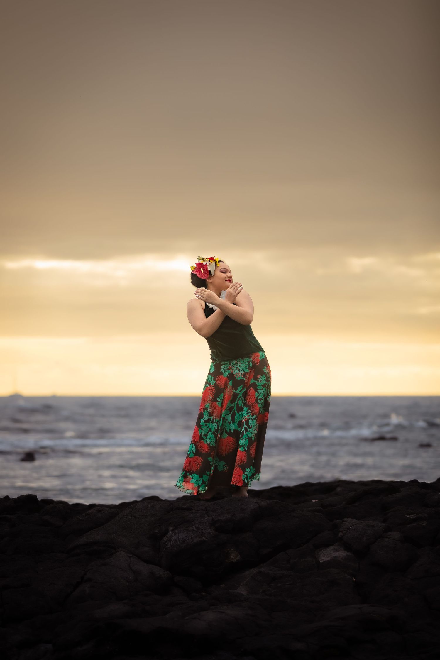 A person in a floral skirt poses dramatically on black rocks at sunset by the ocean.