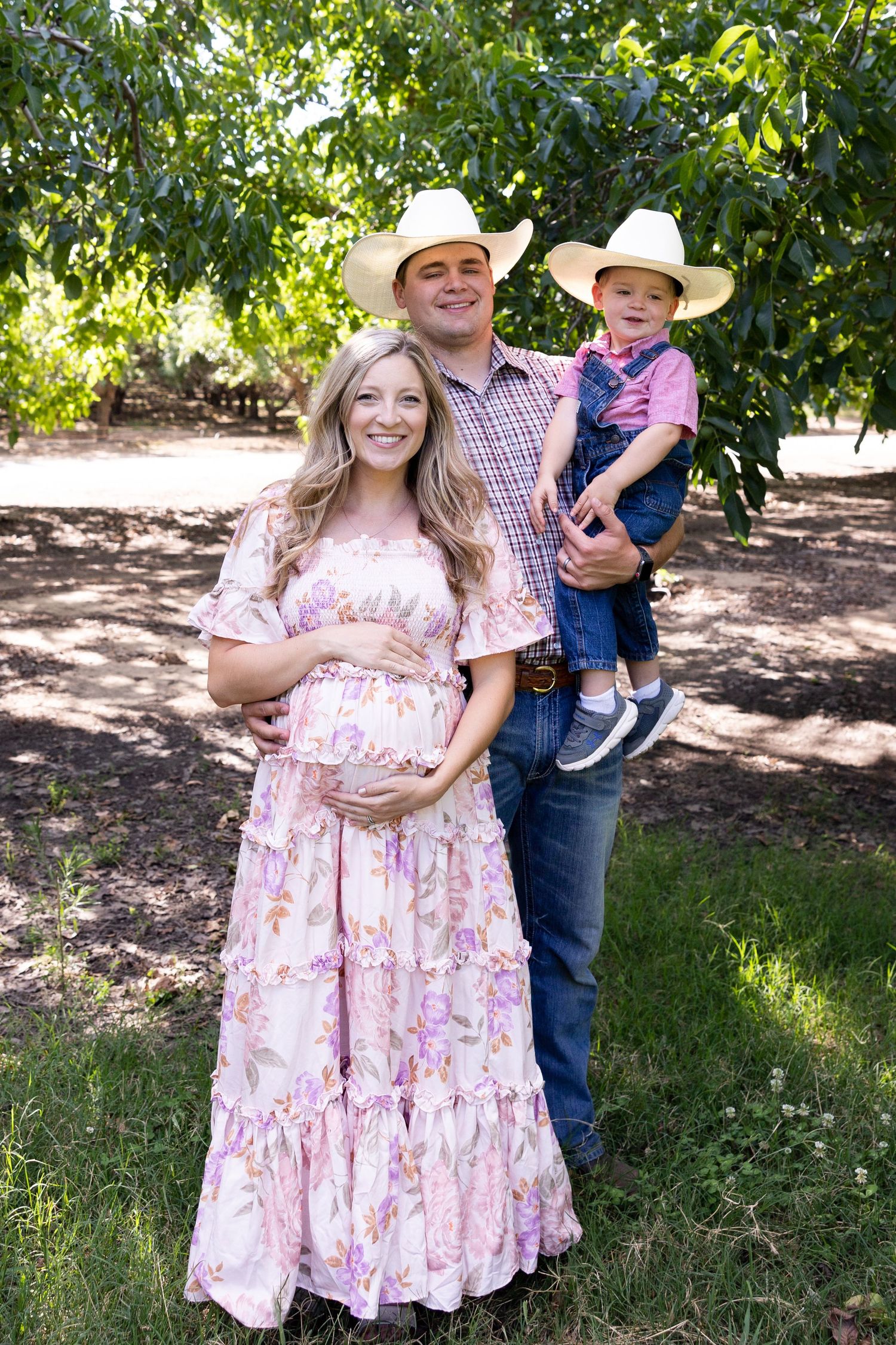 Family during maternity session. Woman is bolding belly in Orchard in Orland, California. Boys have on cowboy hats